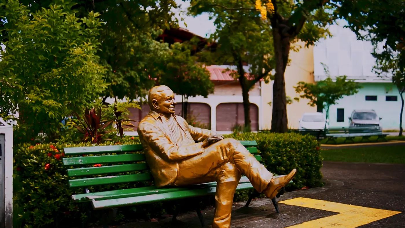 Estátua em tom dourado de um homem sentado em um banco verde de praça, cercado por plantas