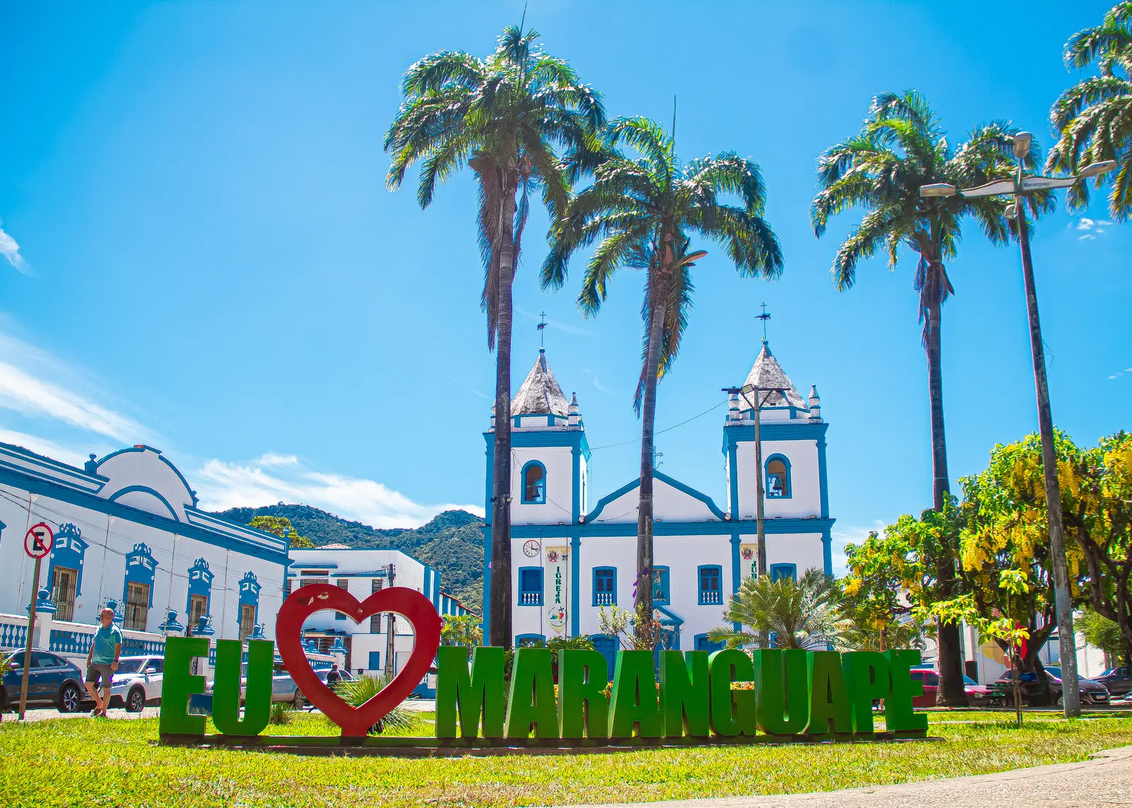 Letreiro colorido com a frase Eu amo Maranguape e um coração vermelho, em praça com palmeiras e igreja branca e azul ao fundo