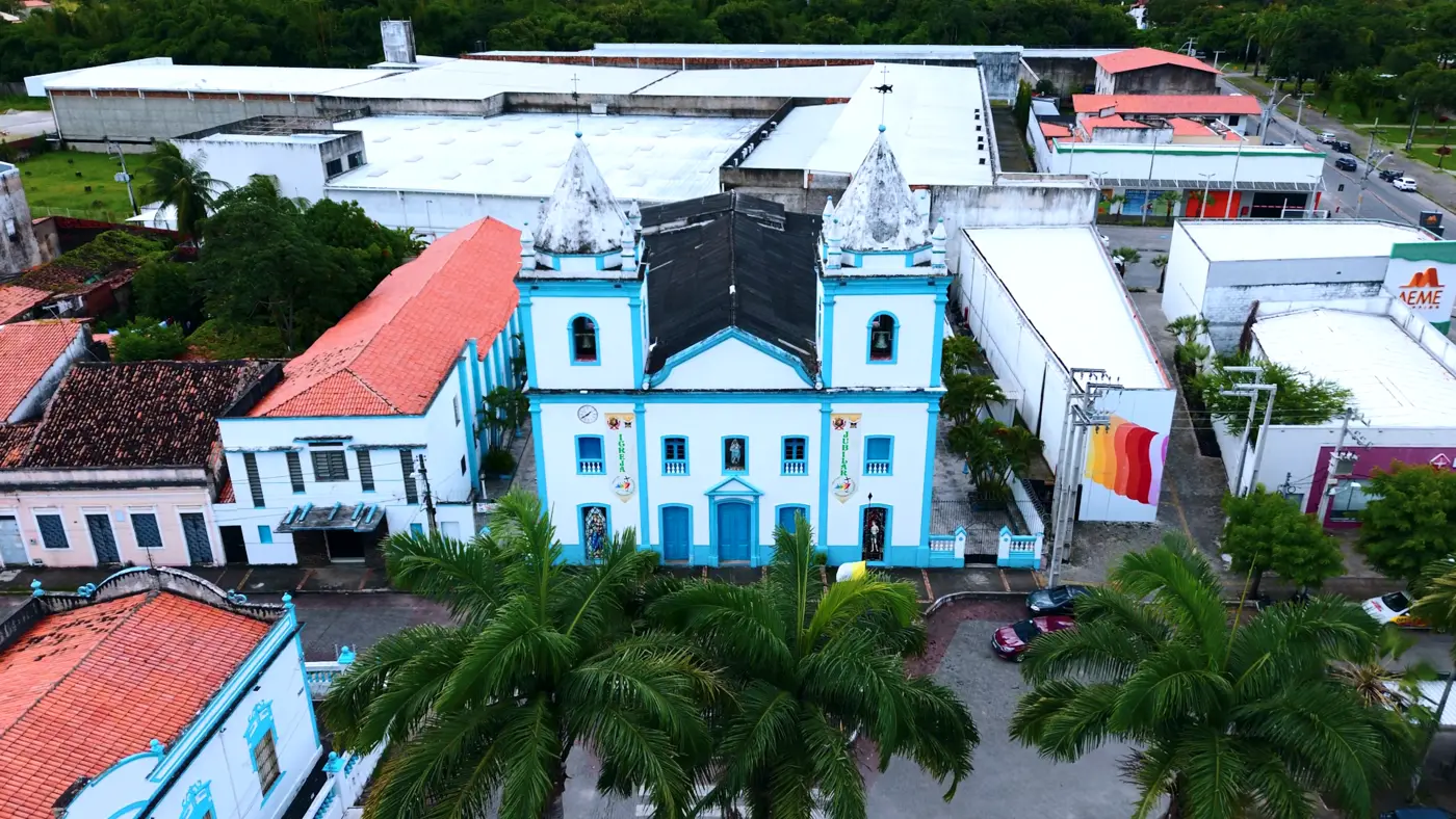 Vista aérea de uma igreja branca com detalhes azuis e duas torres, entre palmeiras e construções do centro da cidade
