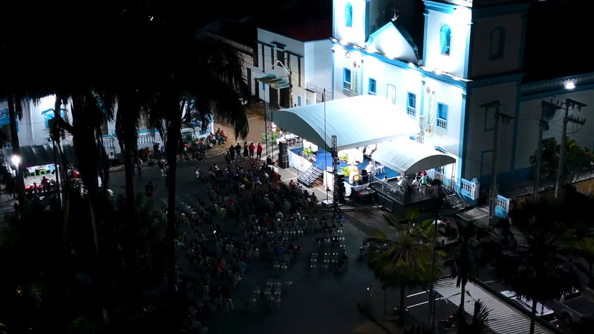Igreja branca e azul iluminada à noite, com público reunido na praça em frente