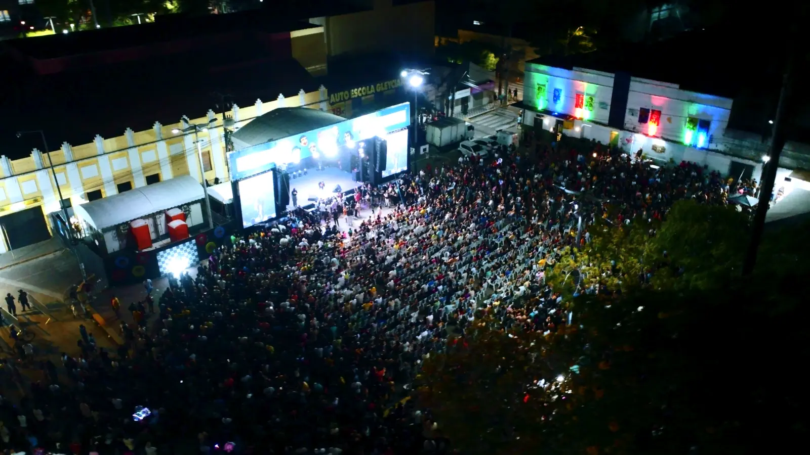 Vista aérea noturna de uma grande multidão diante de um palco iluminado em praça da cidade
