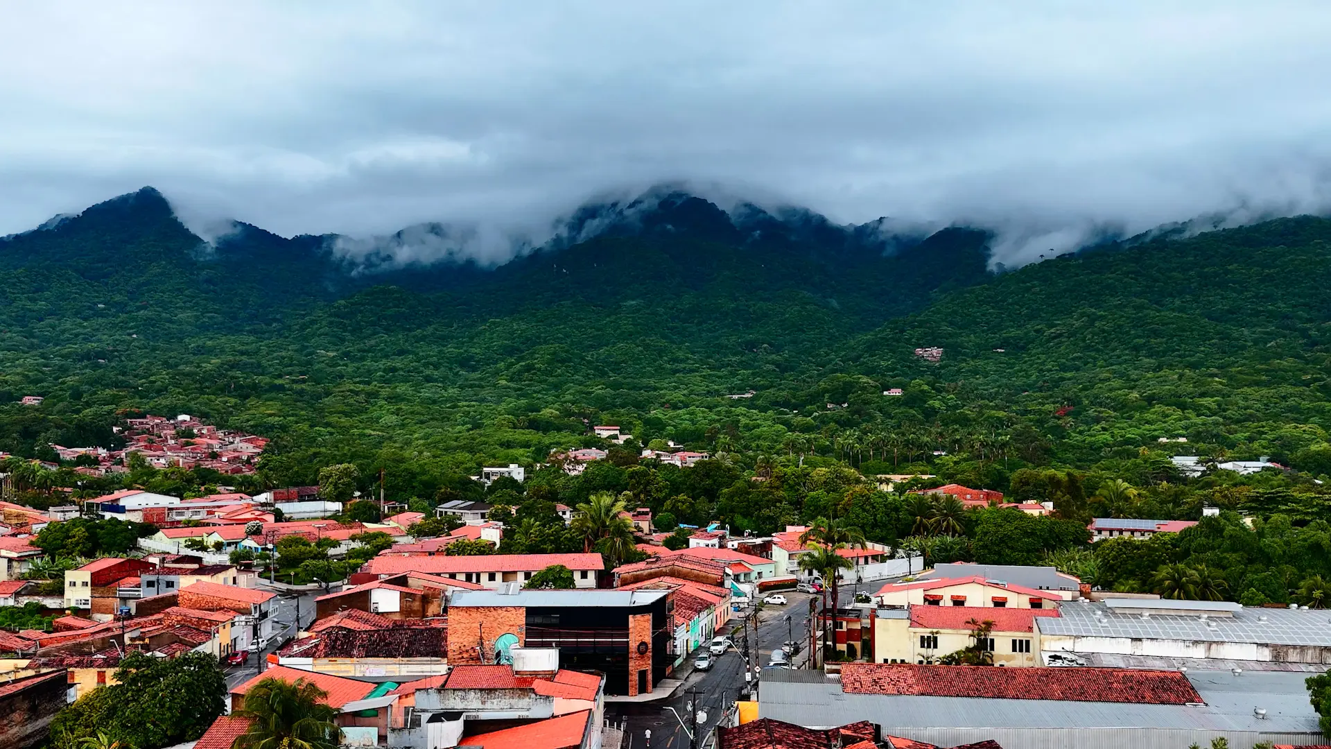 Vista aérea de Maranguape com casas de telhado cerâmico em primeiro plano e a serra coberta de mata e nuvens ao fundo